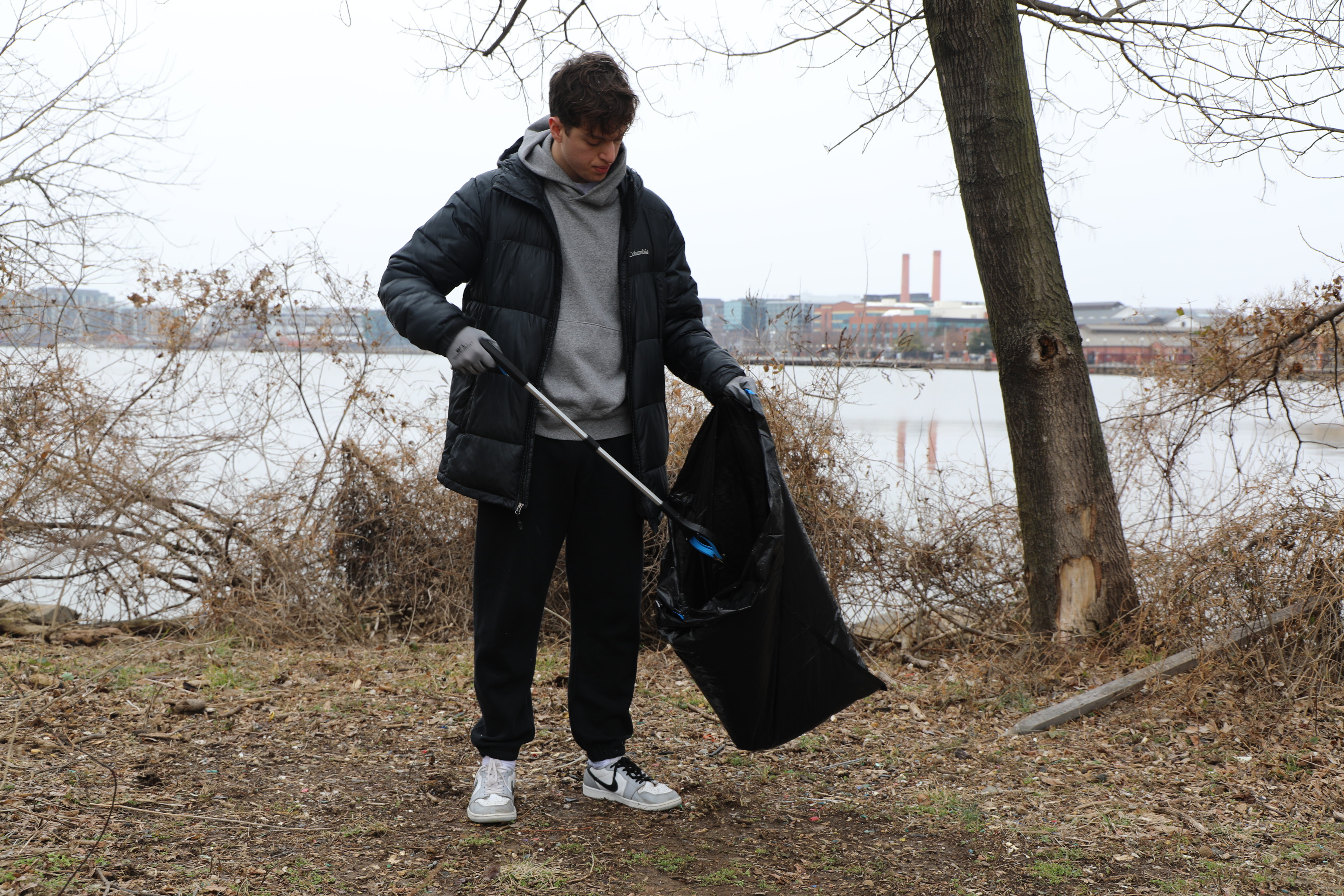 A young man stands outdoors on a patch of dry leaves and sparse grass, beside a large body of water. He is wearing a black puffer jacket over a grey hooded sweatshirt, black sweatpants, and white sneakers. He has on grey gloves and is holding a black trash bag in his left hand, while using a long grabber tool with his right hand to pick up waste from the ground and place it in the bag. To his right, a tree trunk is visible, and in the background across the water, there are industrial buildings with smokestacks under a grey sky. The trees around him are bare.