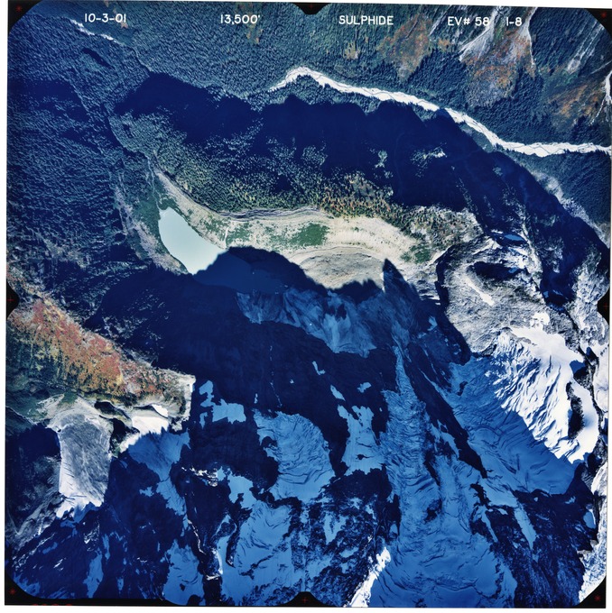 Color aerial photograph of glacier on mountain peak with forested valleys below.