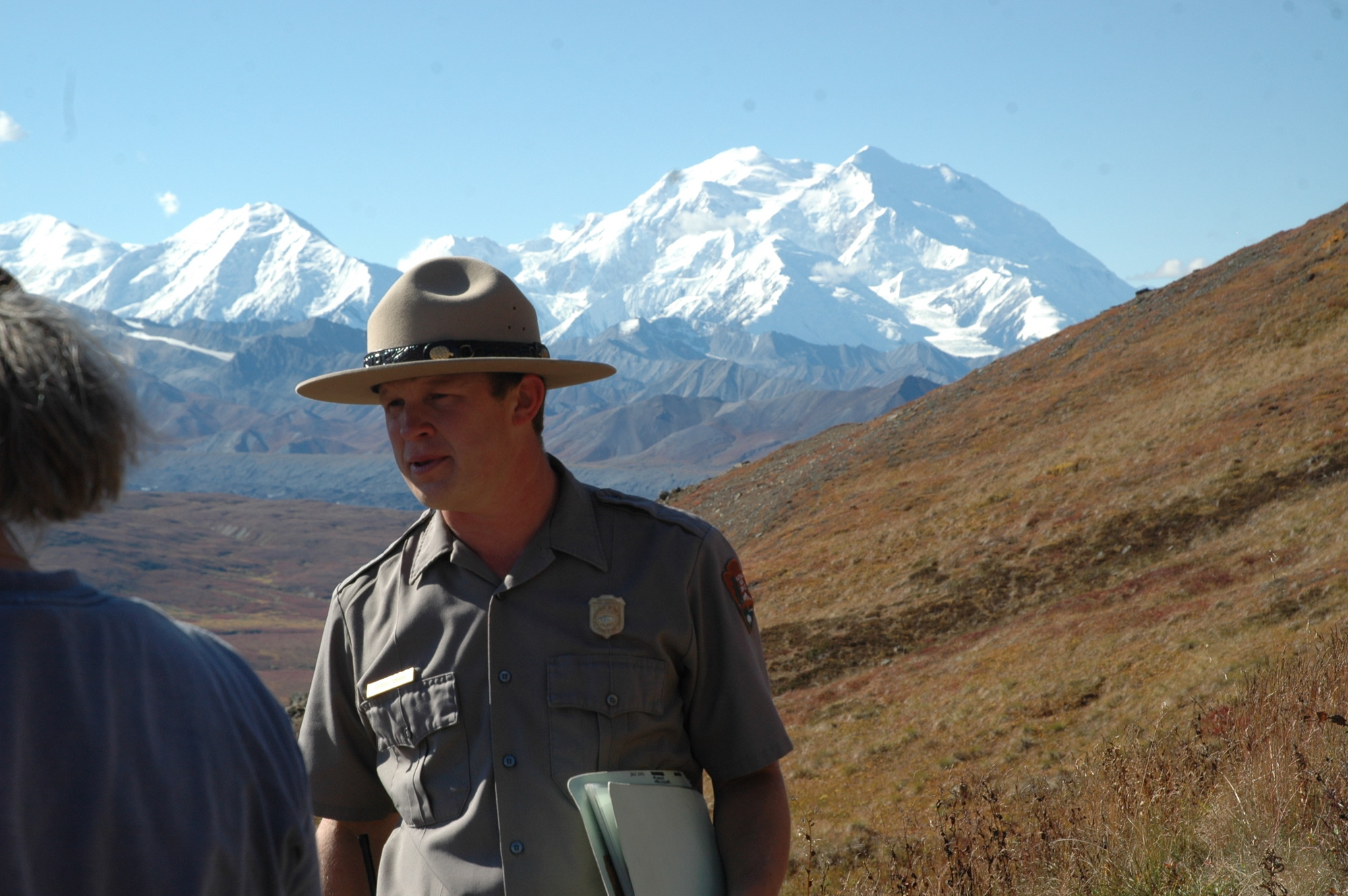 ranger and visitor on a mountain looking at a distant, snowy peak