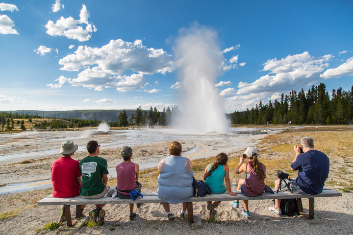 Adults and children sit on a bench looking out over a geyser which erupts at an angle to the left.
