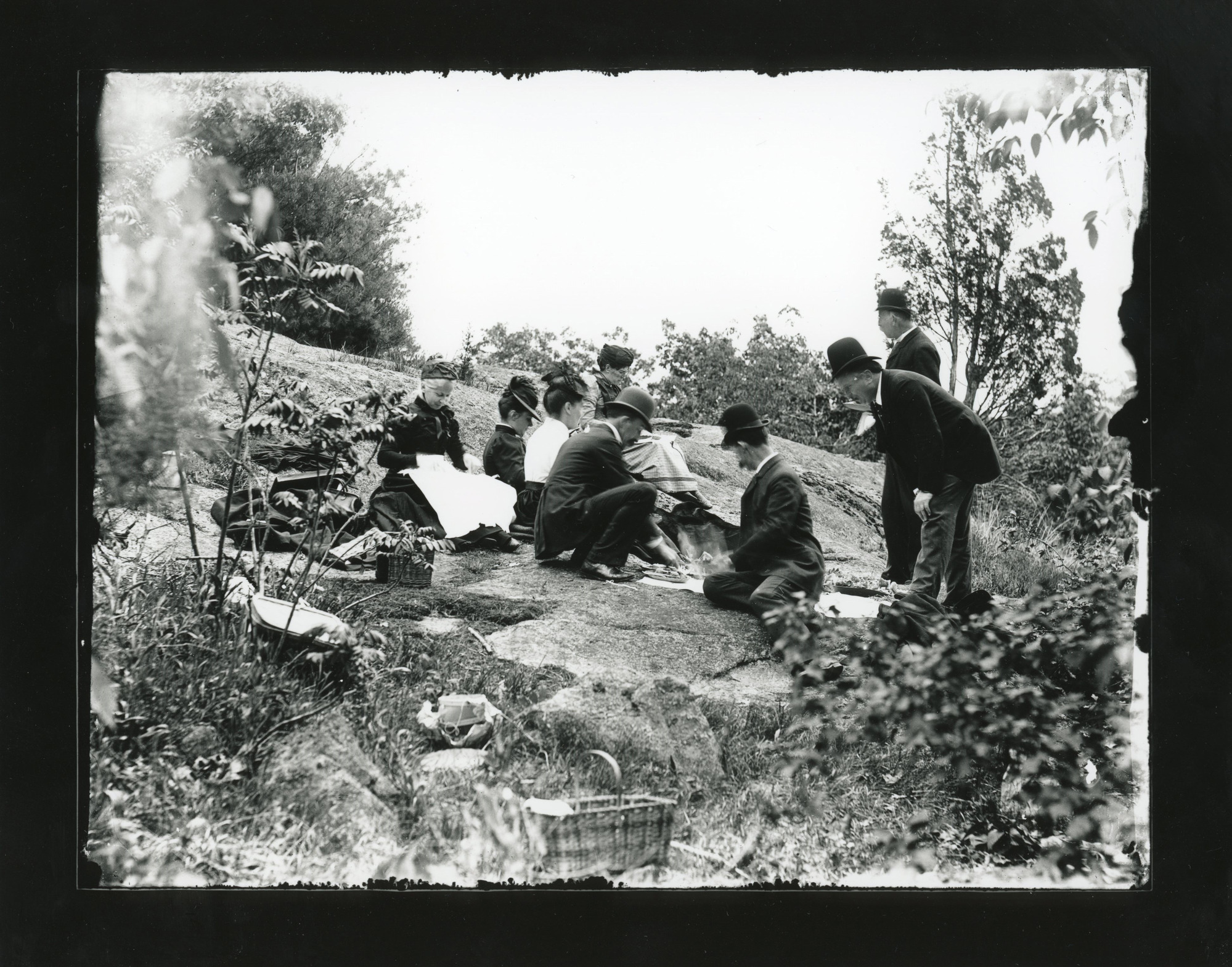Four white men and four women in full formal dress picnic on a large flat rock in the woods.