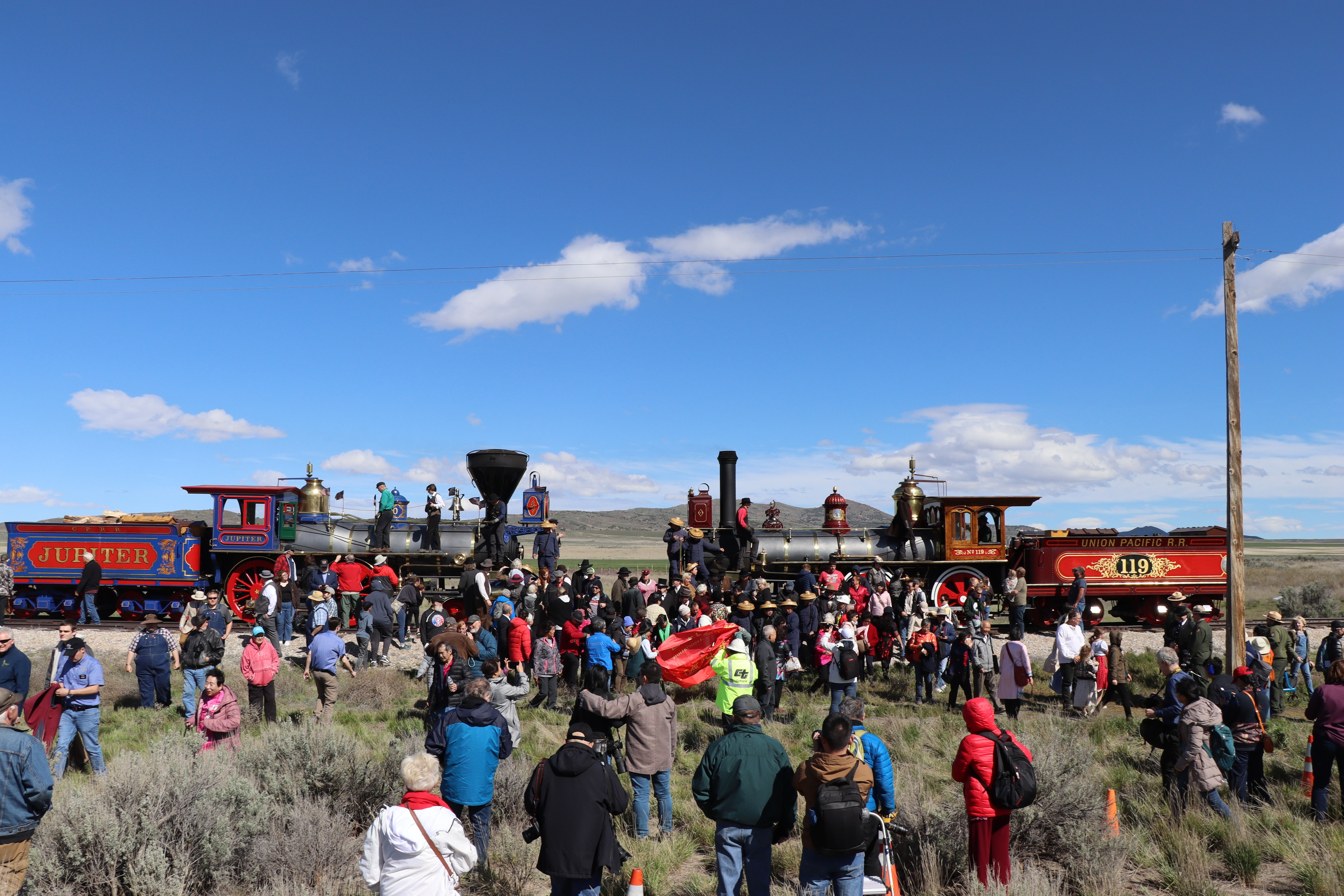 Men and women, some in 1860s period dress and some in traditional Chinese clothing, gather in front of and atop two steam locomotives in a recreation of Andrew J. Russell's famous photograph. The two train engineers, hanging off their respective locomotives in the center of the frame, hold out glass bottles. 
