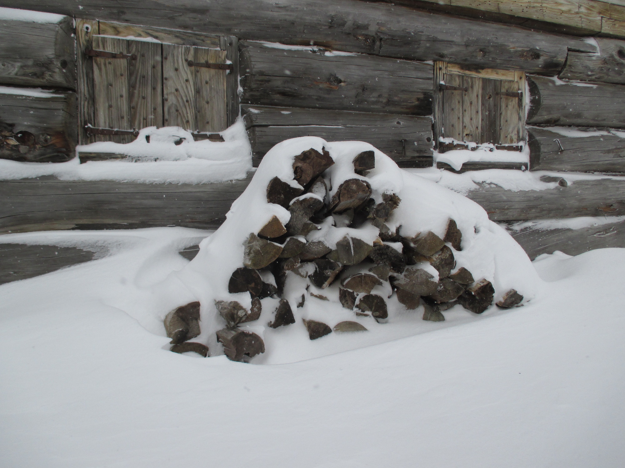 A triangular pile of wood in front of a wooden building is buried in snow.