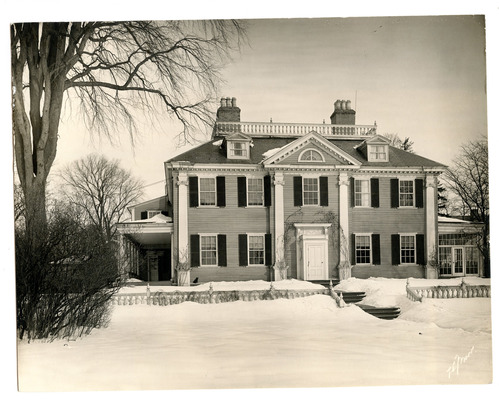 Black and white photo of facade of Georgian mansion covered in snow, surrounded by bare trees.