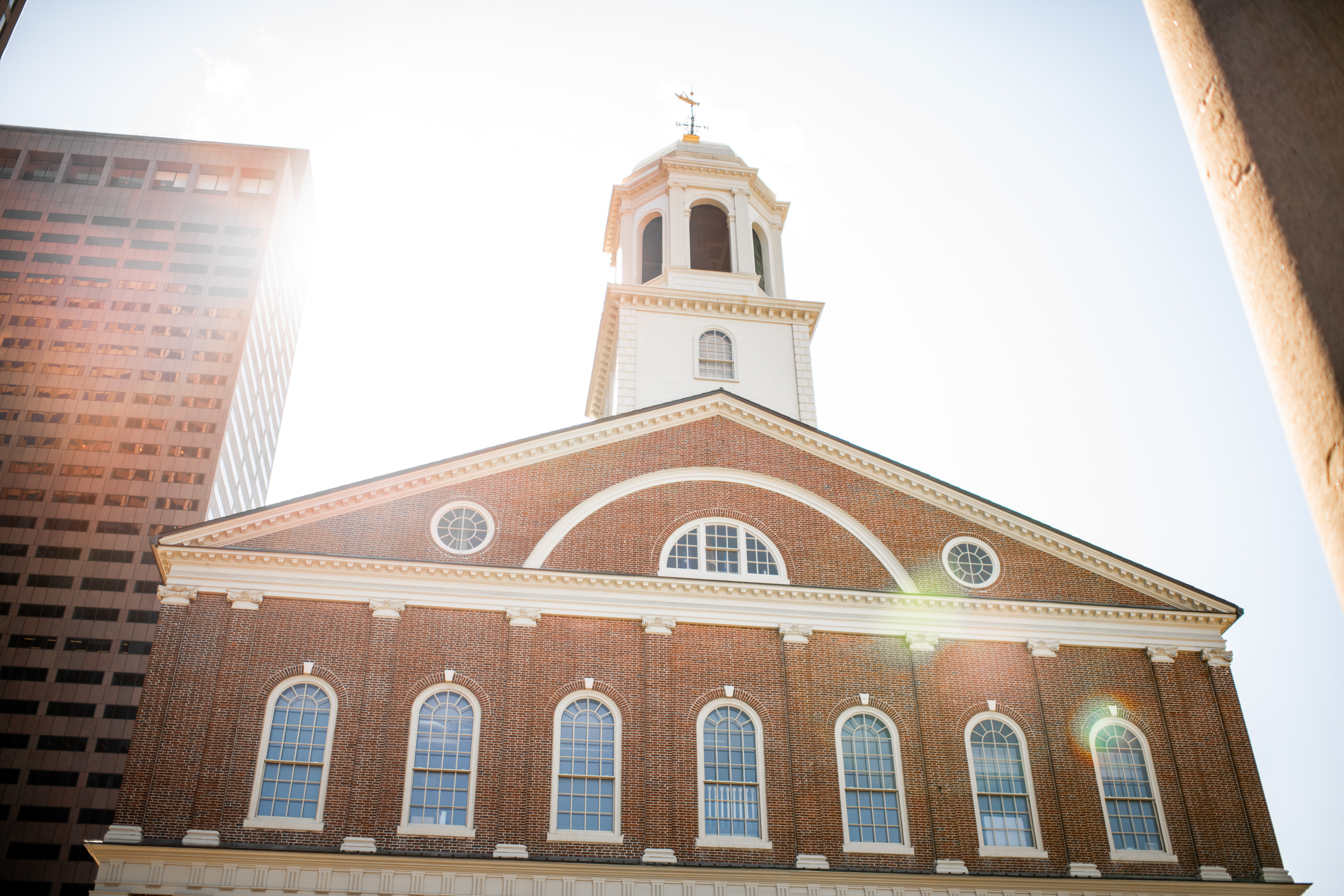View looking up at the top floors of a federal-style brick building known as Faneuil Hall. The building has rows of arched windows on each floor and is topped by a pediment with two circular windows on either side of a half-circle window. The building has a cupola with a grasshopper weathervane.