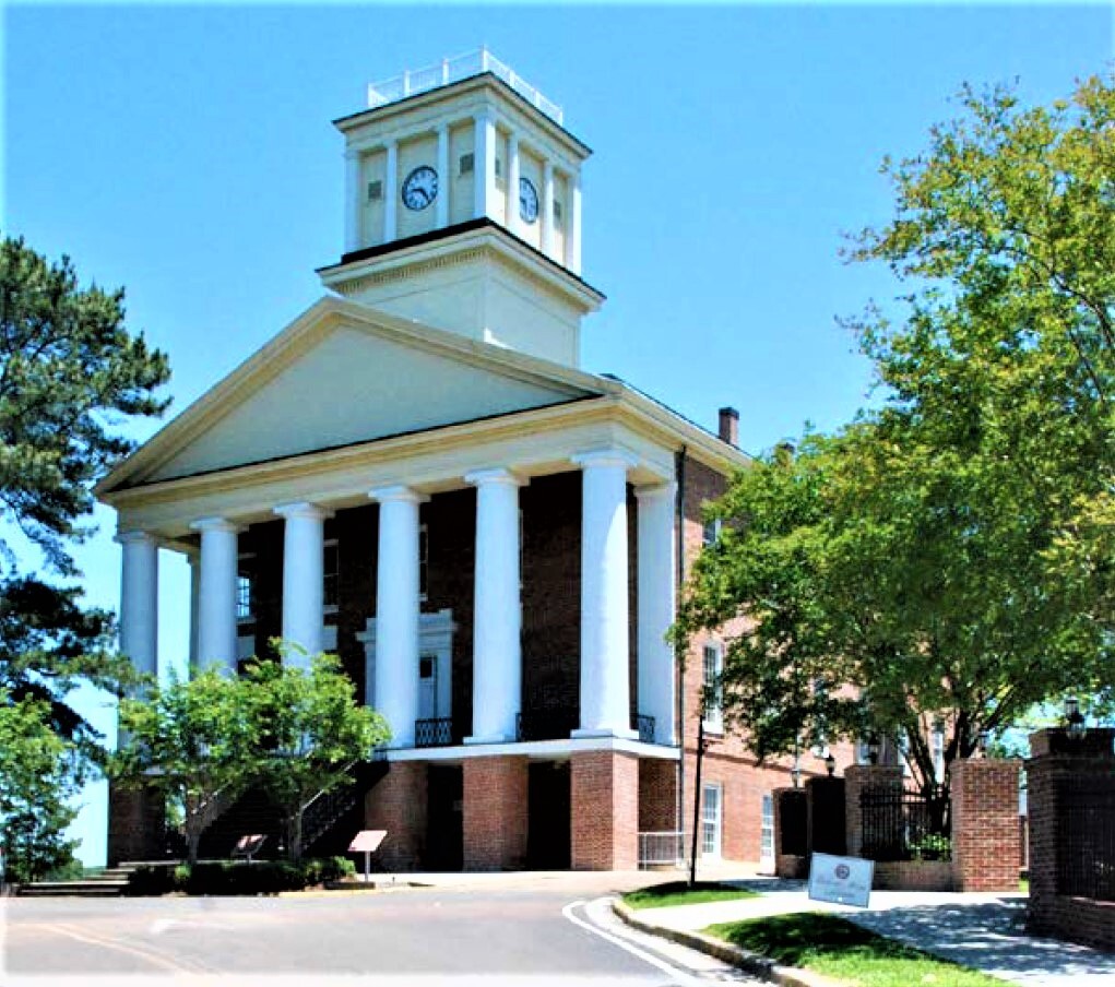 Color photo of greek revival style chapel building. The exterior is brick with white plaster columns at the front, topped by a triangular plinth. Above the plinth is a large square cupola that serves as a clock tower. Walkways, grass, and trees surround the building.