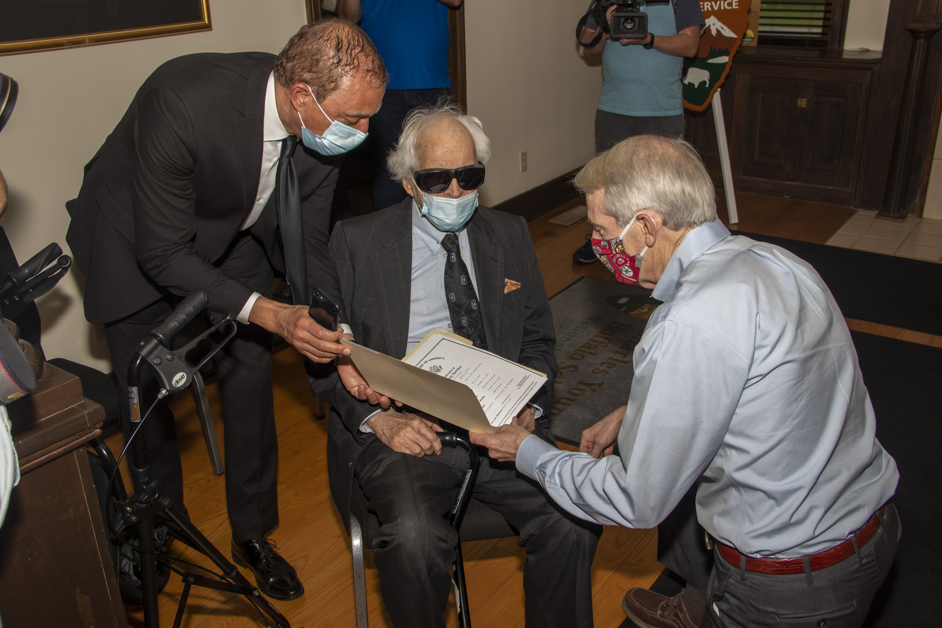 A man in a blue shirt and red mask looks at a folder with documents inside of it as a man in a suit and tie and blue mask holds onto it.