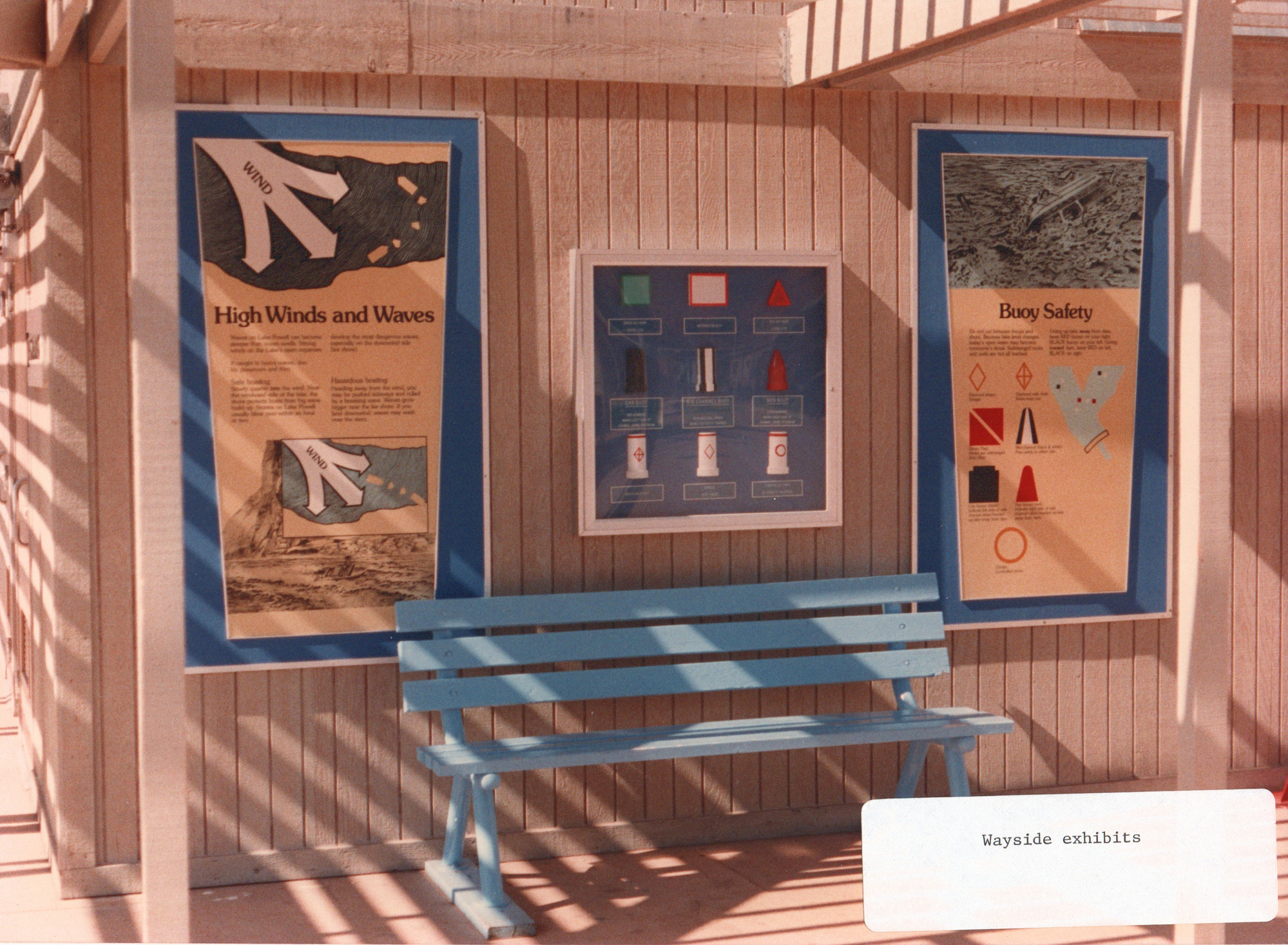 A blue bench sits at the base of three signs describing water safety and aids to navigation. label in the corner of the photo: Wayside exhibits