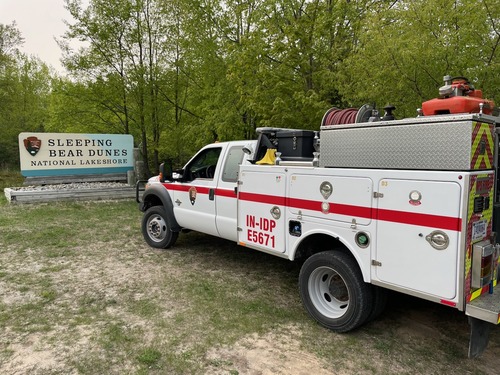 A white fire truck with a red stripe, built on a pick-up truck chassis, parked in front of a large Sleeping Bear Dunes National Lakeshore sign. 