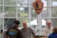Two people in uniforms talk to visitors standing underneath a National Park Service arrowhead. 
