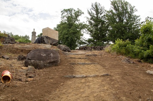 A brown stabilized aggregate path with natural stone water bars in it sits between topsoil and runs up a hill between large rocks towards a monument surrounded by boards at the top of the slope, with the top of another monument and trees in the background. 