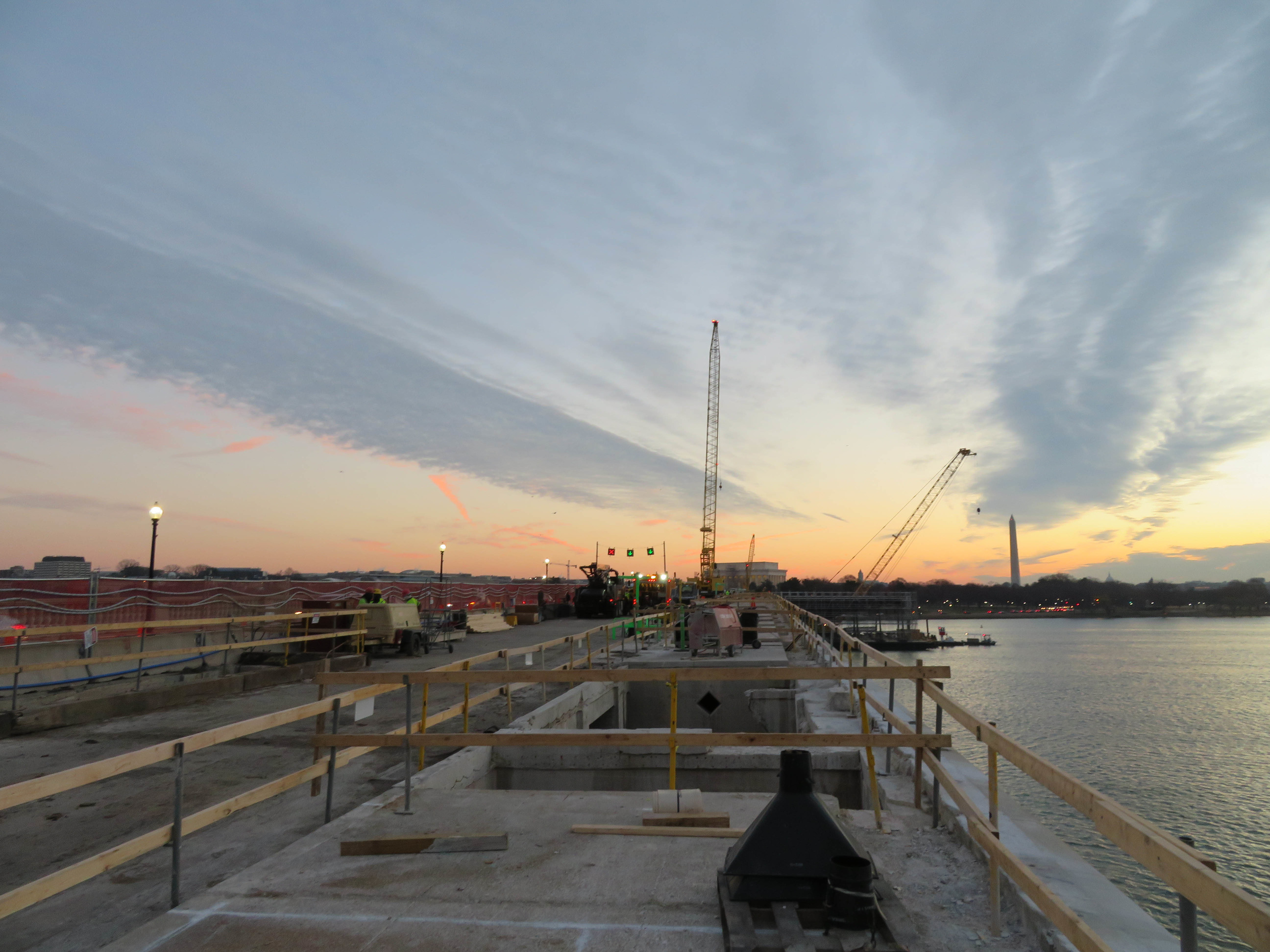 The sun sets as construction on the bridge takes place. Two large cranes are in the distance. 