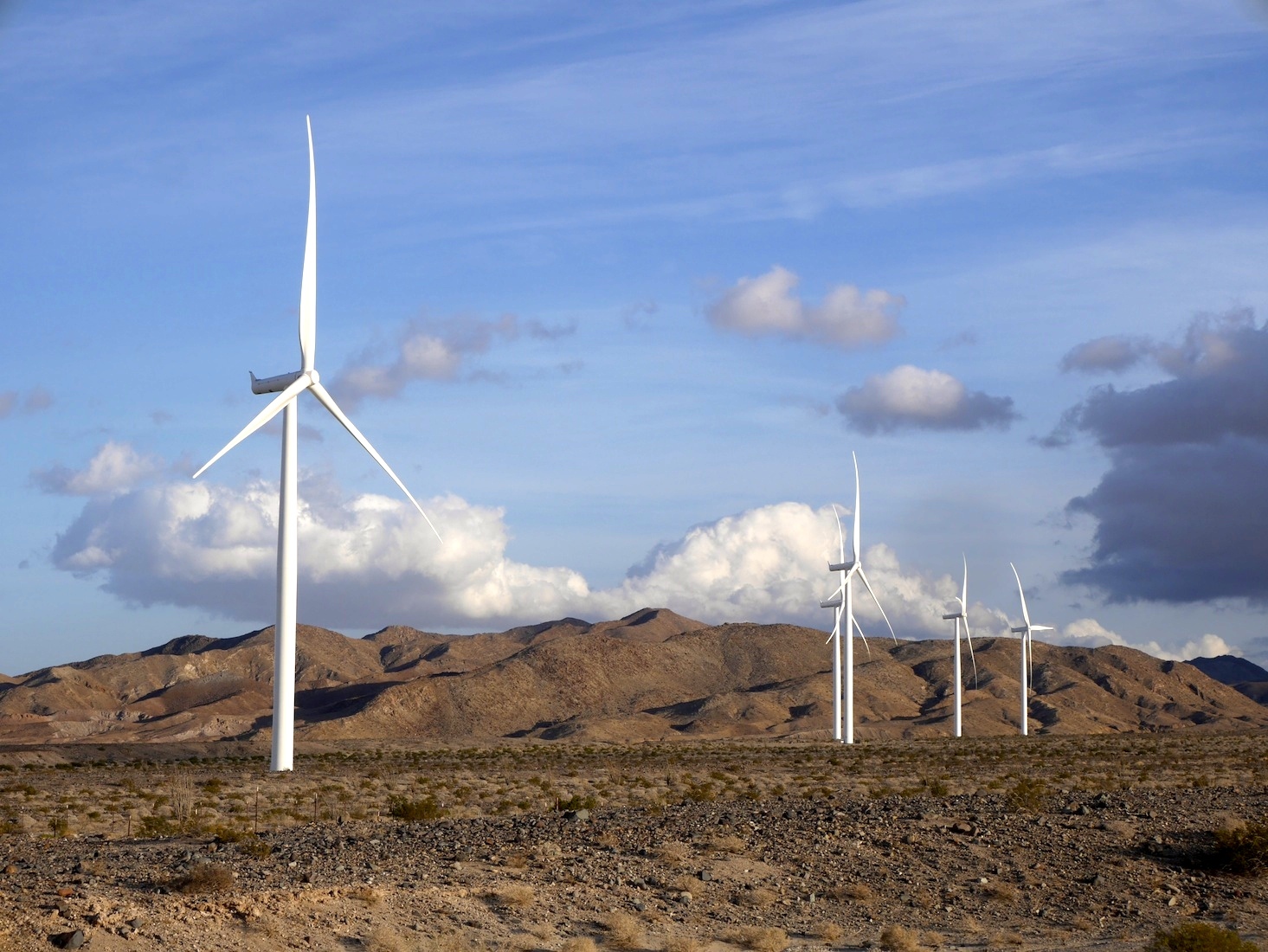 Five wind turbines stand on a rocky landscape with golden hills and clouds