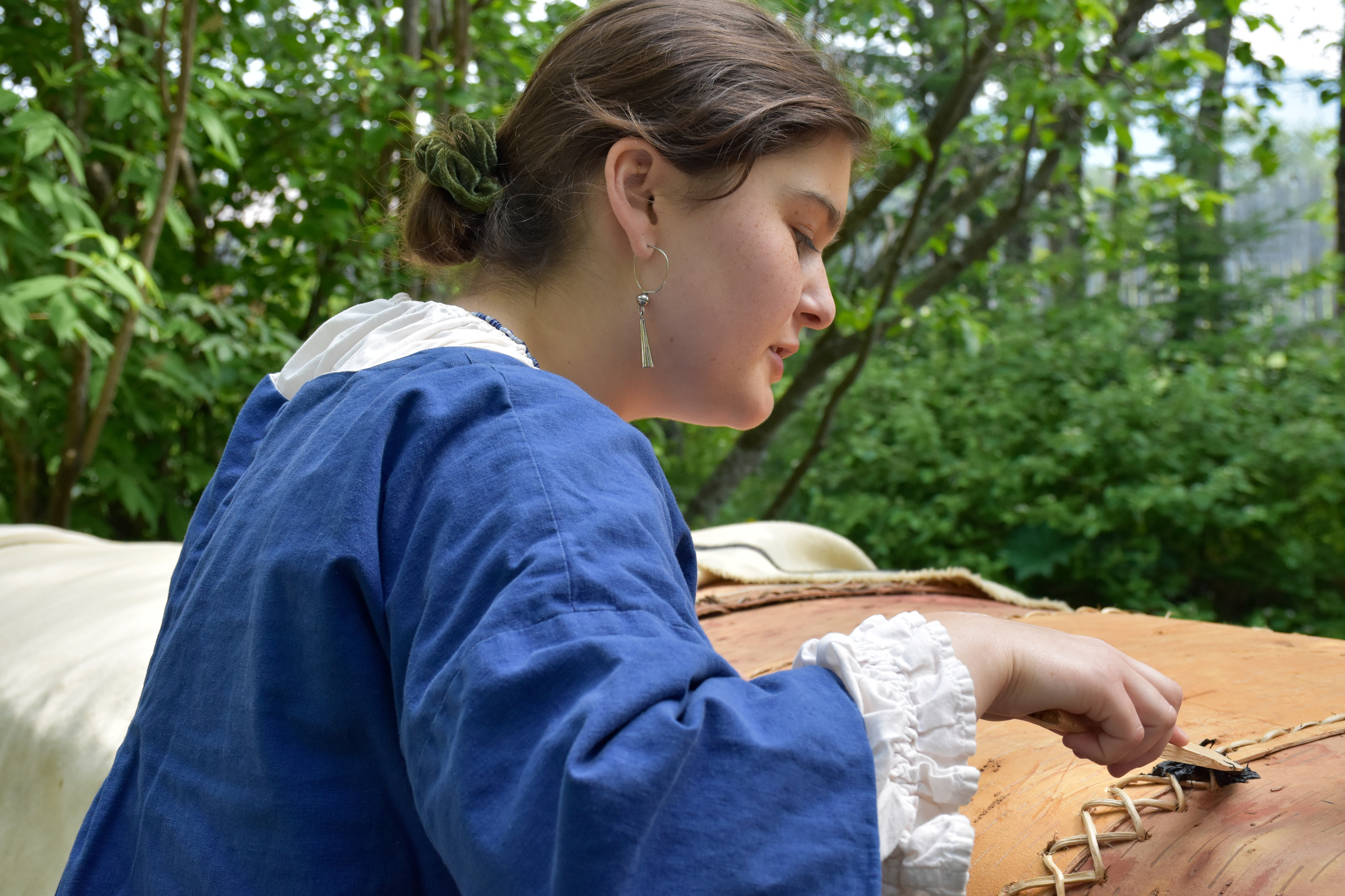 A person wearing a historic blue jacket with white shirt cuffs and neckline showing applies a black substance to coarse stitching on a curved bark surface.