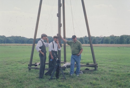 Three men stand by a wooden catapult. 