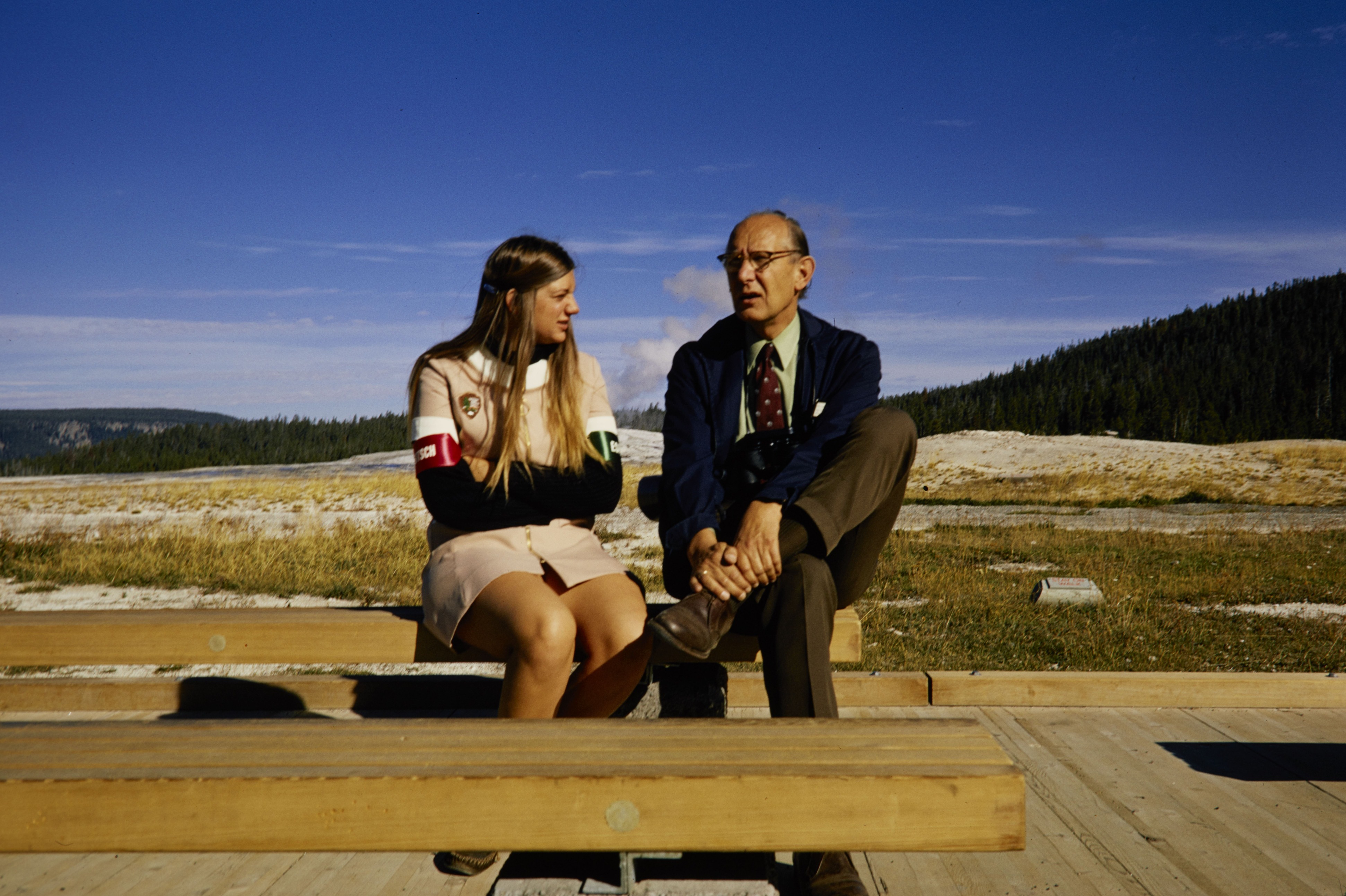 Woman in a beige NPS uniform dress sits on a bench talking a man in a suit.