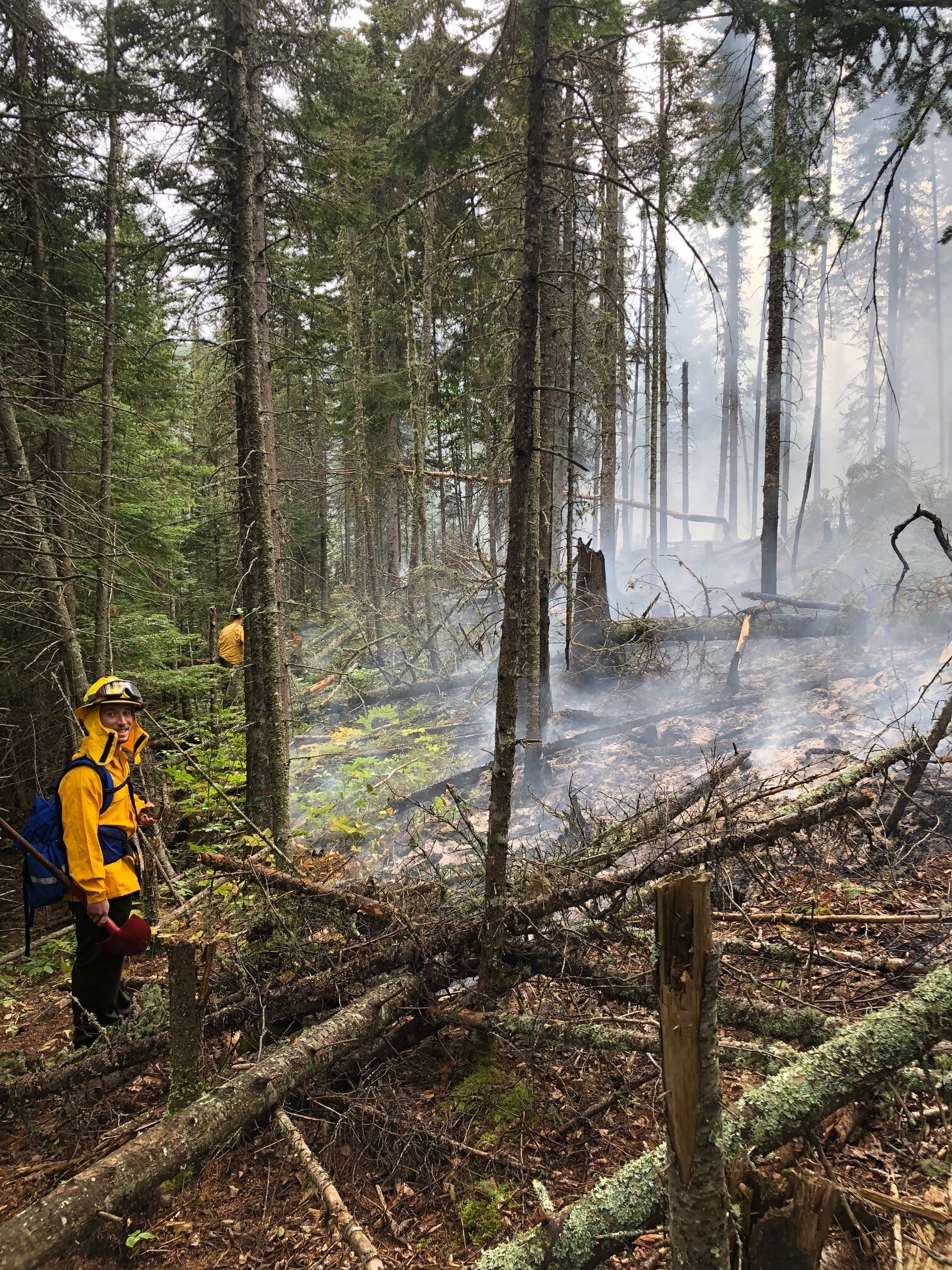 A wildland firefighter wearing a yellow shirt and blue fire pack smiles for the camera while holding a red shovel.