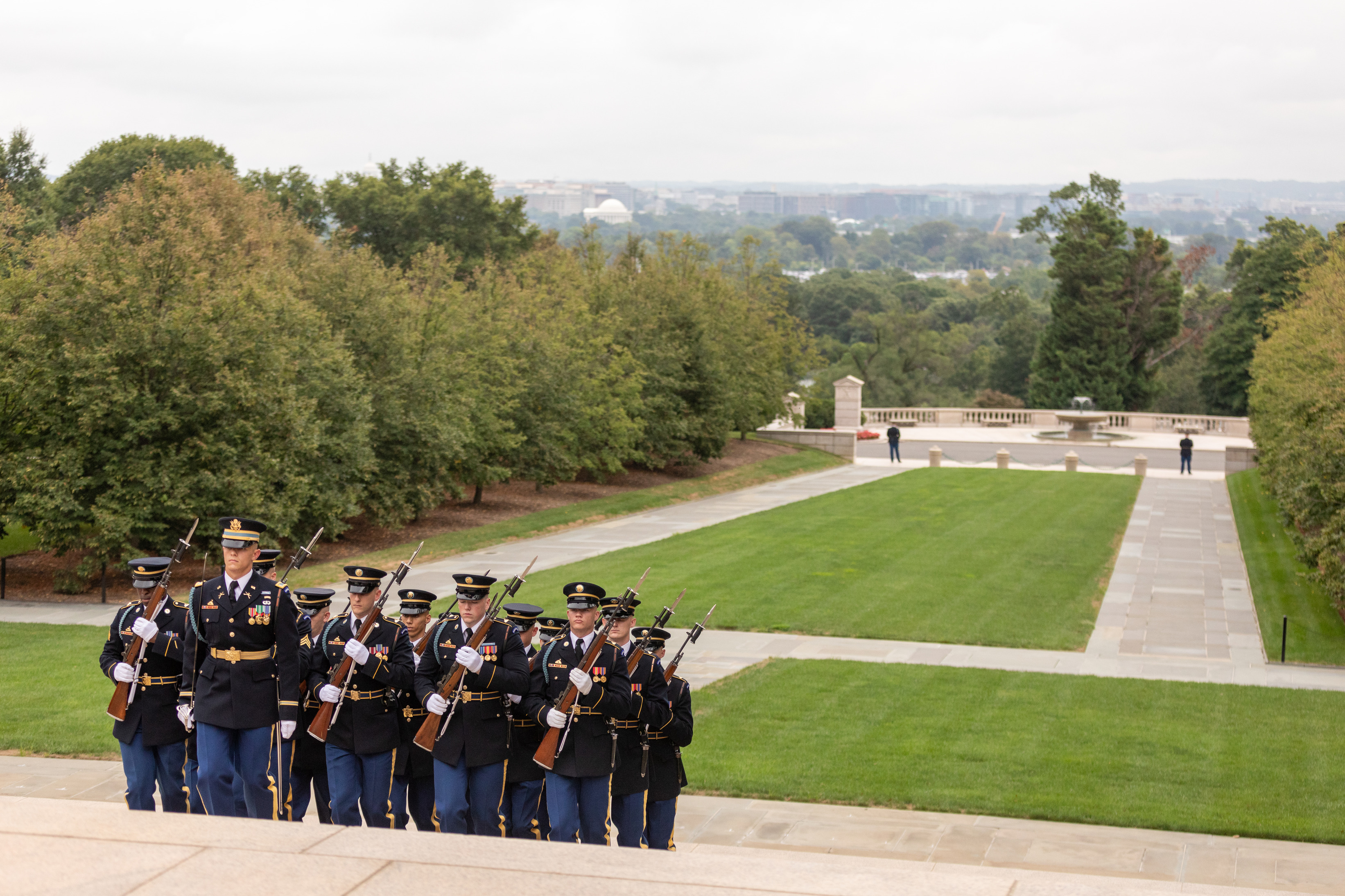 A group of soldiers march with rifles in their hands. The Washington DC skyline and the Thomas Jefferson Memorial loom in the distance.