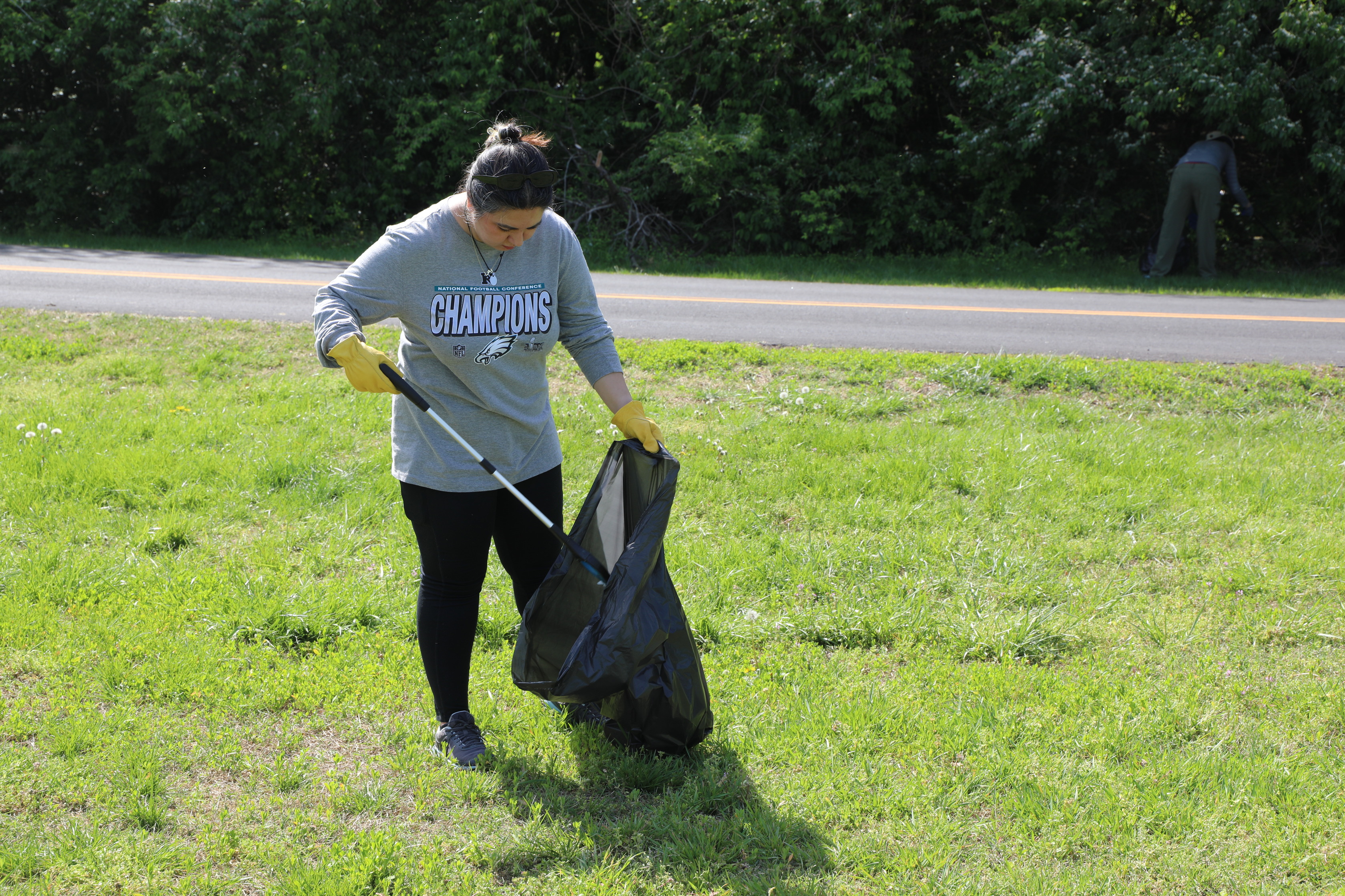 A person with dark hair tied back in a bun, wearing a grey long-sleeved shirt with "CHAMPIONS" written on it, black pants, and yellow gloves, stands in a grassy field. They are using a litter picker to put trash into a black trash bag held open in front of them. In the background, a road runs parallel to a dense line of green bushes and trees. Another person is partially visible in the far-right background, also appearing to be picking up litter. The day is bright and sunny.