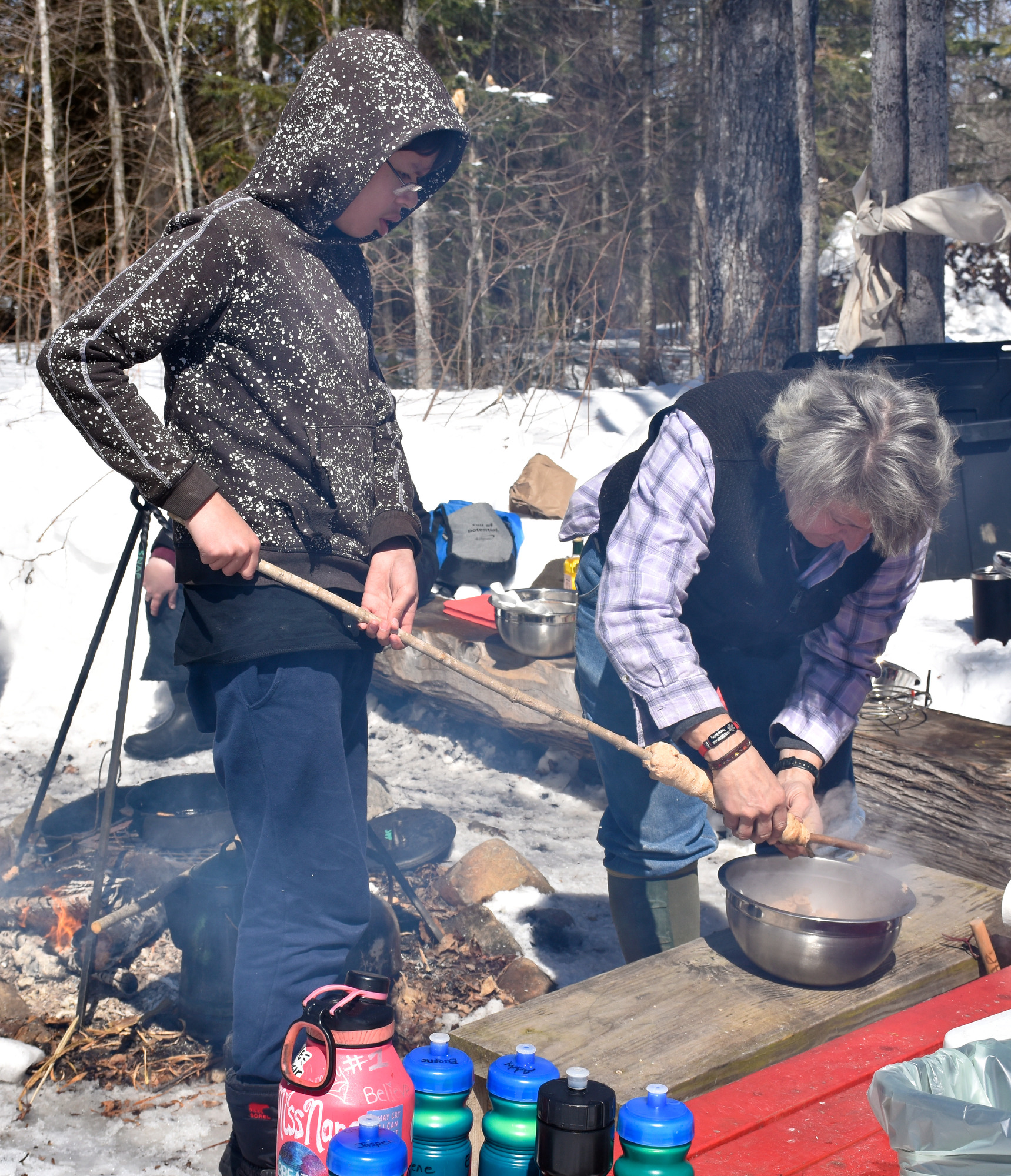 A student holds a long stick with bread wrapped around it while a community member peels off the baked bread into a waiting bowl.