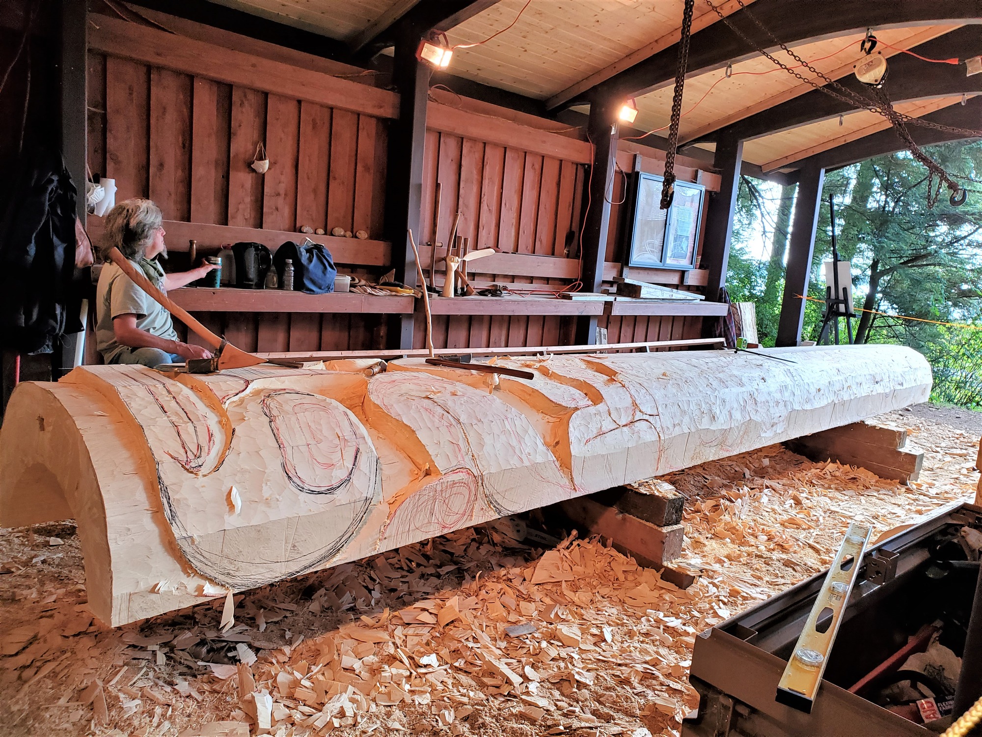 A man in a covered shed, next to a horizontail, partially carved pole. There are red and black lines depicting ovoids and curved shapes. There are woodchips on the ground. An adze is sitting on top of the pole next to the man.