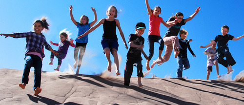 Ten kids jumping off a sand dune beneath a blue sky
