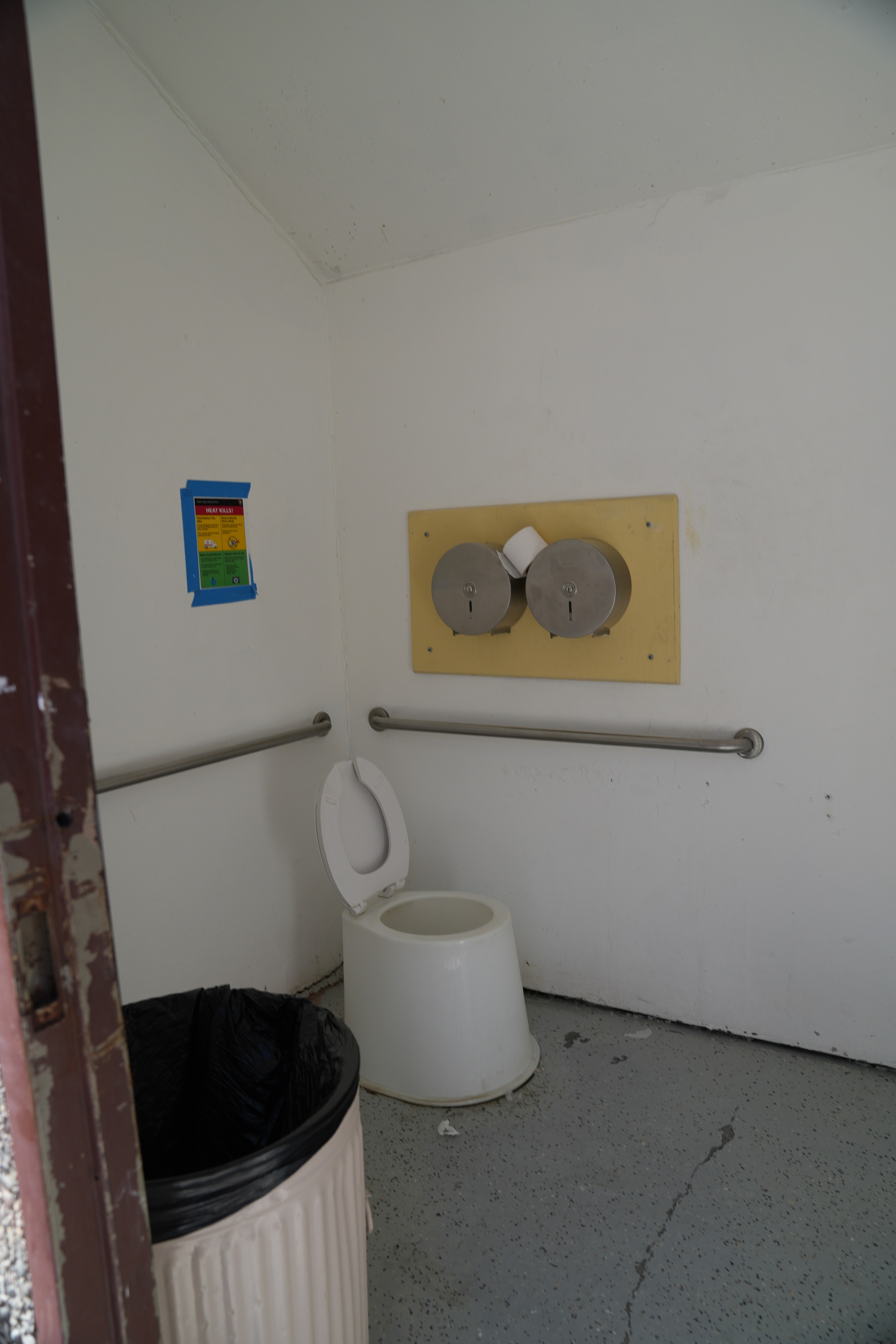 Badwater Basin bathroom interior. A vault toilet with grab bars on the side and back end. A trash can is inside the restroom. The floor is made of concrete and it is even.