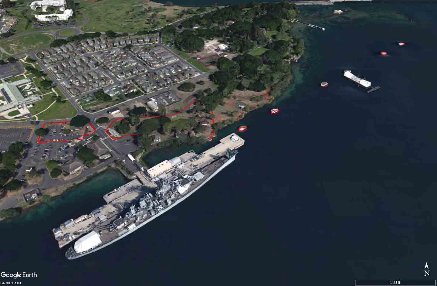 Aerial view of the shoreline at Pearl Harbor. The cultural landscape boundary is near the shore 
 where a battleship is docked along a pier, shown using a red outline in the photo.