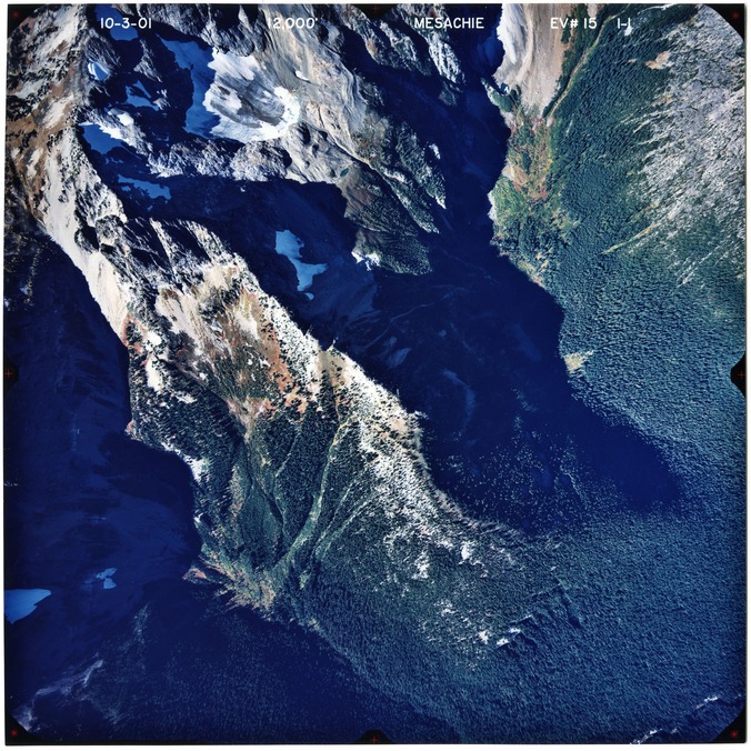 Color aerial photograph of glaciated mountain peak with forested valleys below.