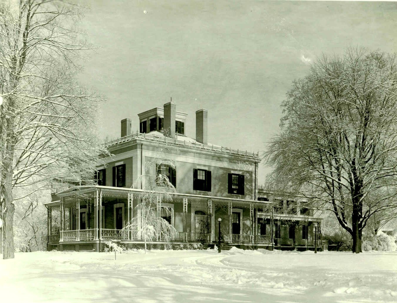 Black and white photo of a large brick house with a wraparound porch. A large tree is on either side of the house with one small tree in front of it. The ground and trees are covered in snow. Tire tracks can be seen in the snow leading up to the house.