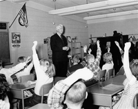 Man standing in the front of a school classroom surrounded by students sitting at desks. 