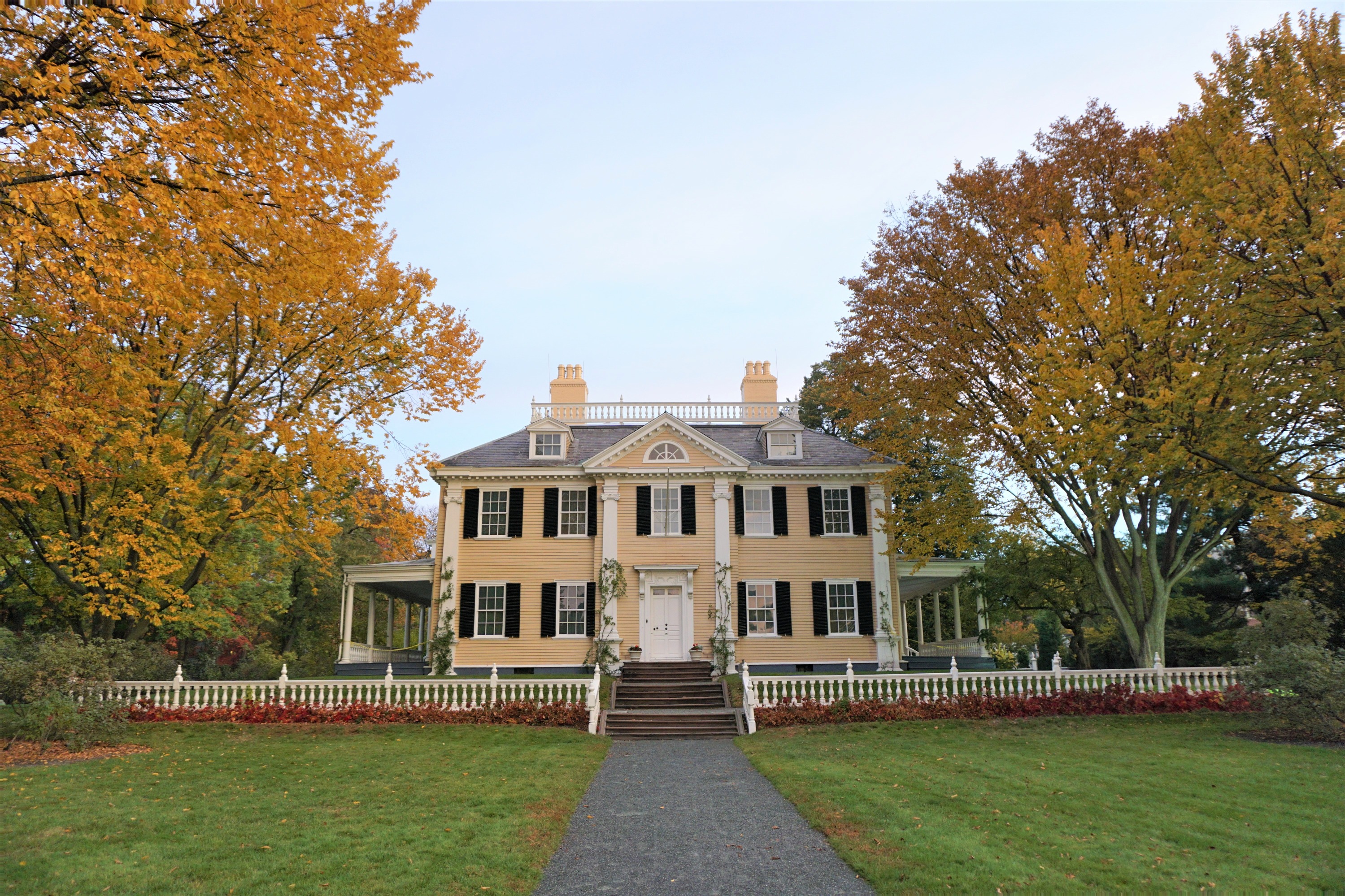 facade of three-story symmetrical yellow Georgian house with large green front lawn, flanked by golden and green elm trees
