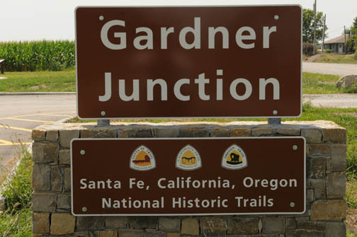 A sign titled with "Gardner Junction" on it beside a road and trees in the background.