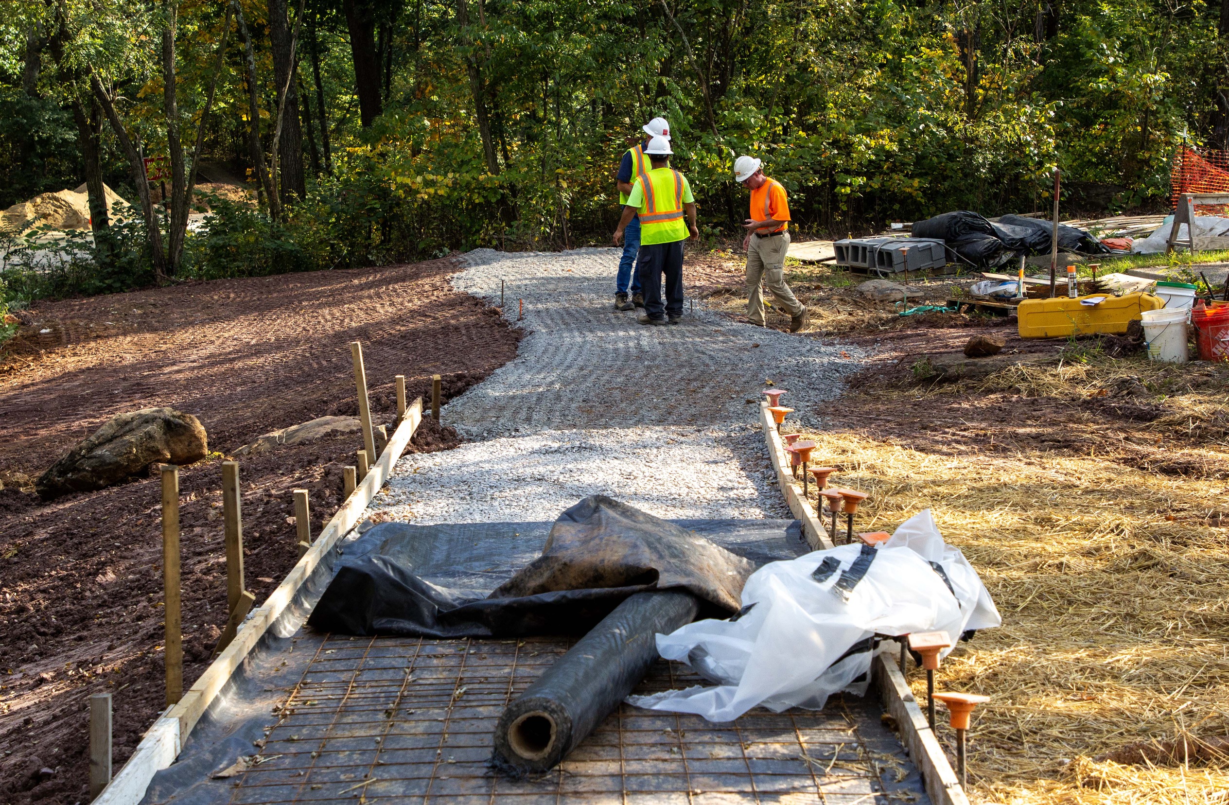 A sidewalk is in various states of preparation. Near the camera the sides have been staked, black construction fabric has been laid, and wire mesh is laid on top. In the distance, only a gravel foundation has been placed down. Three construction workers in white hard hats and bright yellow and orange safety vests assess the work.