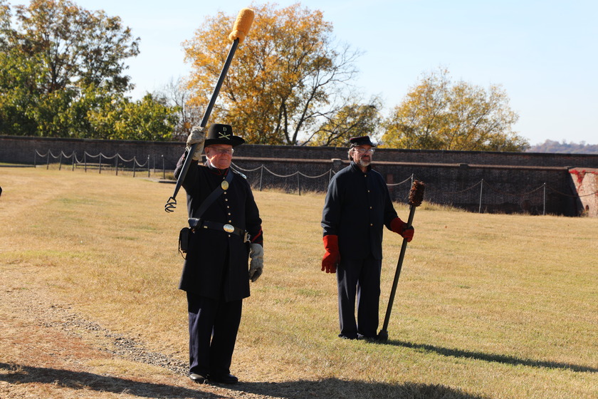 Two men dressed in historical military uniforms stand on a dry, grassy field in front of a stone fort wall. The man on the left is wearing a dark blue Civil War-era Union uniform with a kepi hat, holding a ramrod tool for a cannon raised in his left hand. The man on the right is also in a dark uniform and hat, holding another cannon loading tool, and wearing reddish-brown gloves. In the background, there are trees with autumn foliage under a clear blue sky.