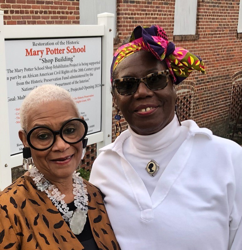 Outdoors, two women stand in front of a large sign next to a brick building. They partially obstruct the sign which reads, in part, Restoration of the Historic Mary Potter School "Shop Building."