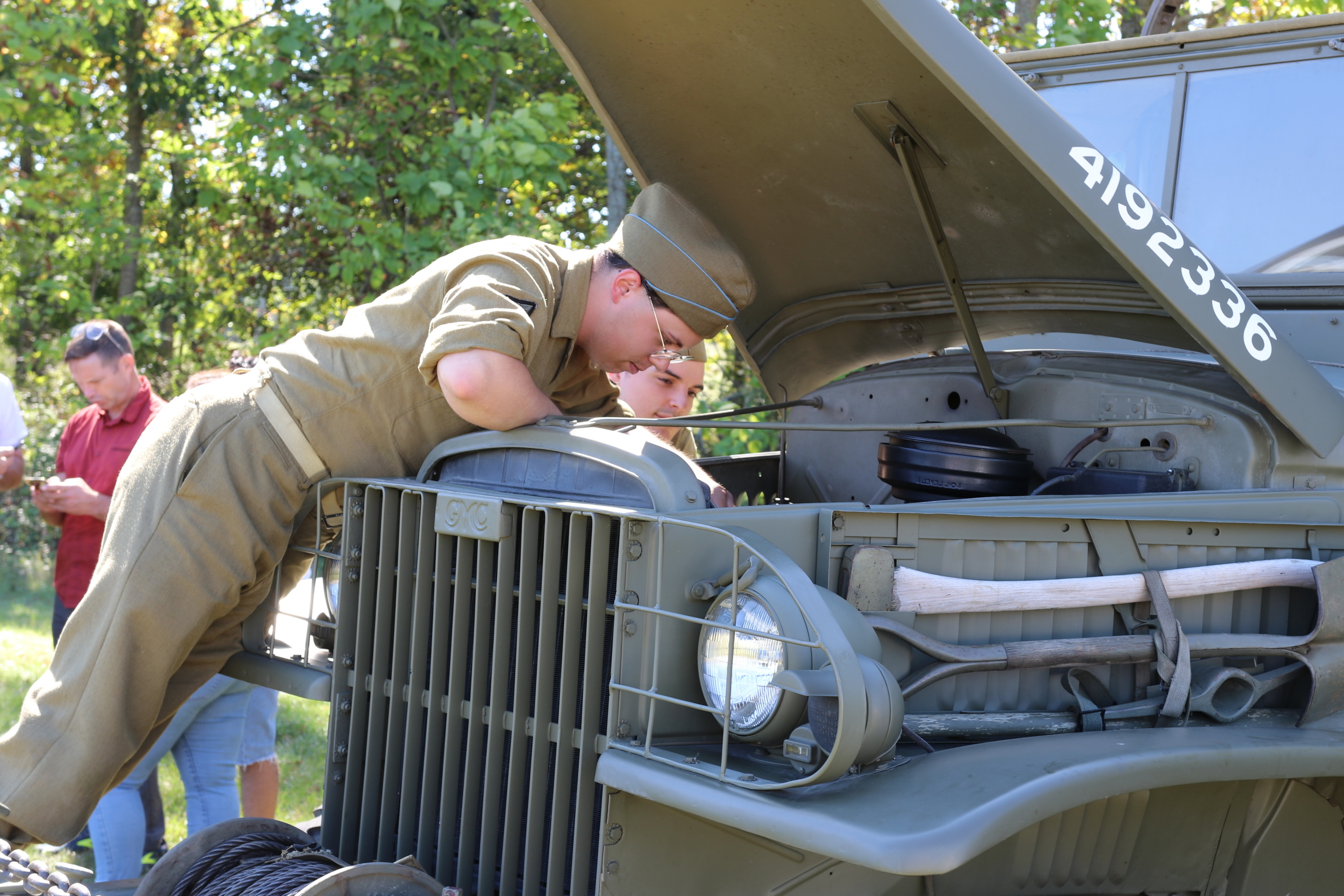 A volunteer wearing a WWII uniform leans under the green hood of a large WWII era vehicle. 