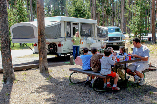 A family sits at a picnic table while the mother stands by a pop up trailer .