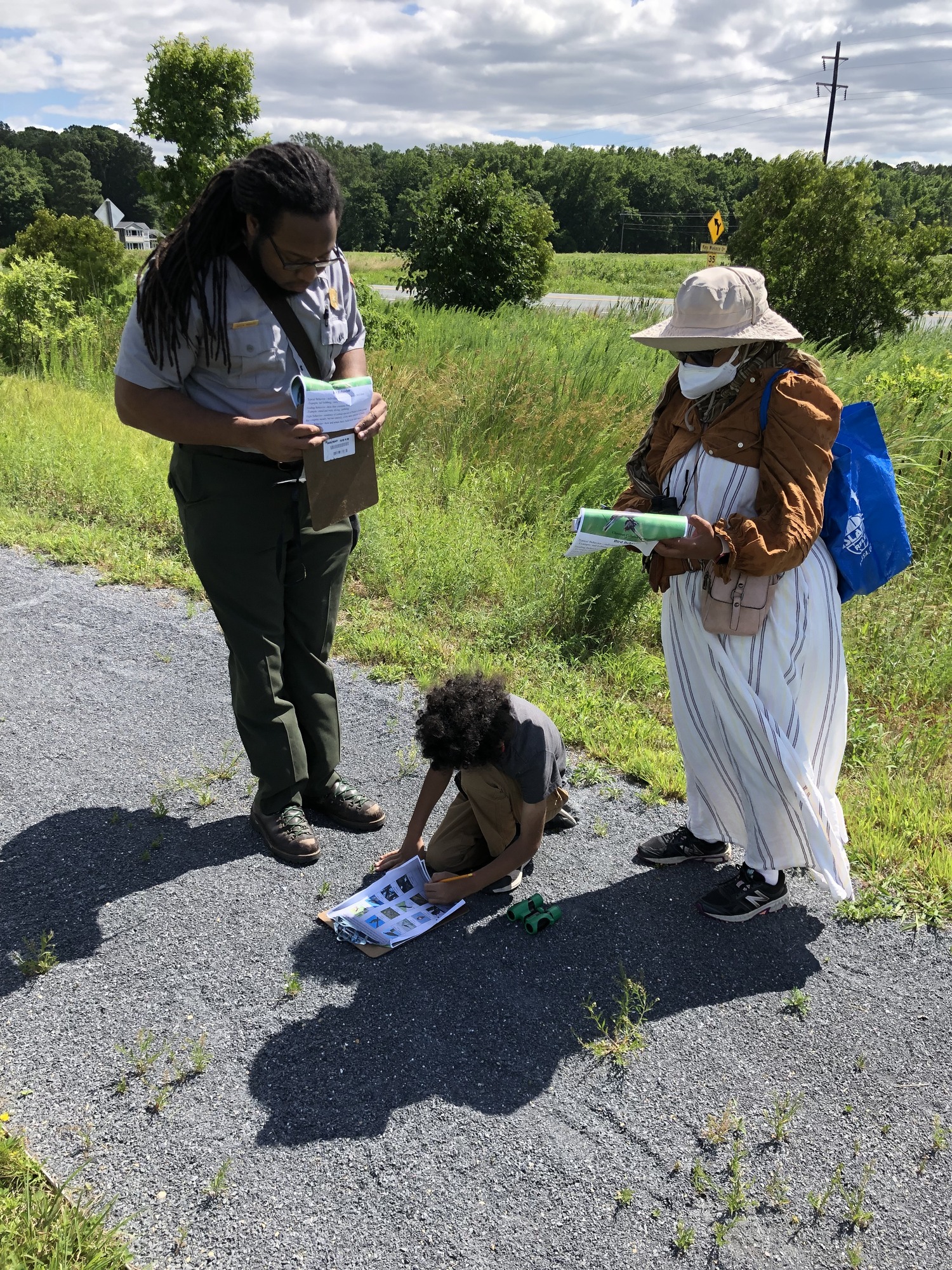 On the left side of the picture, a park ranger holding a clipboard can be seen helping a young boy and his mother with an activity. All three are standing on a blue gravel path with tall grass and trees in the background.