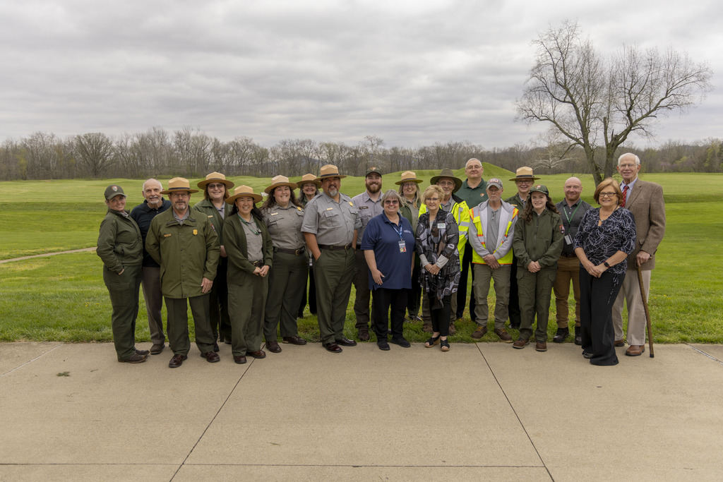Several park rangers in tan flat hats stand alongside several other people in casual clothing pose for a photo in front of green, grass-covered mounds in the background