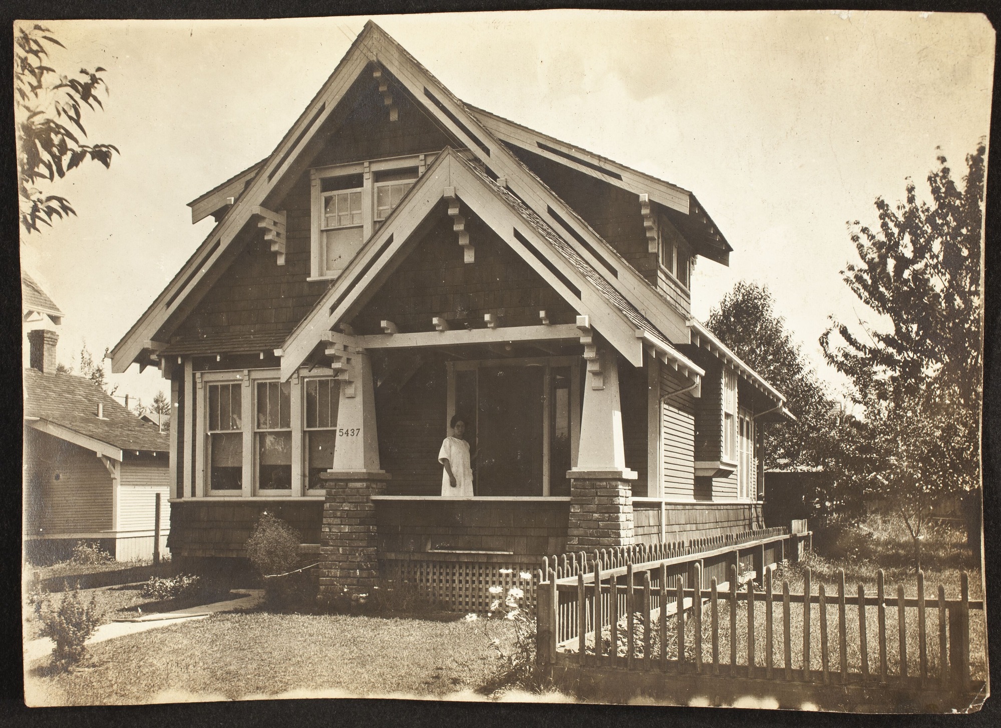 A Craftsman-style house with a woman standing on the front porch.  There is a fenced yard next to the house.
