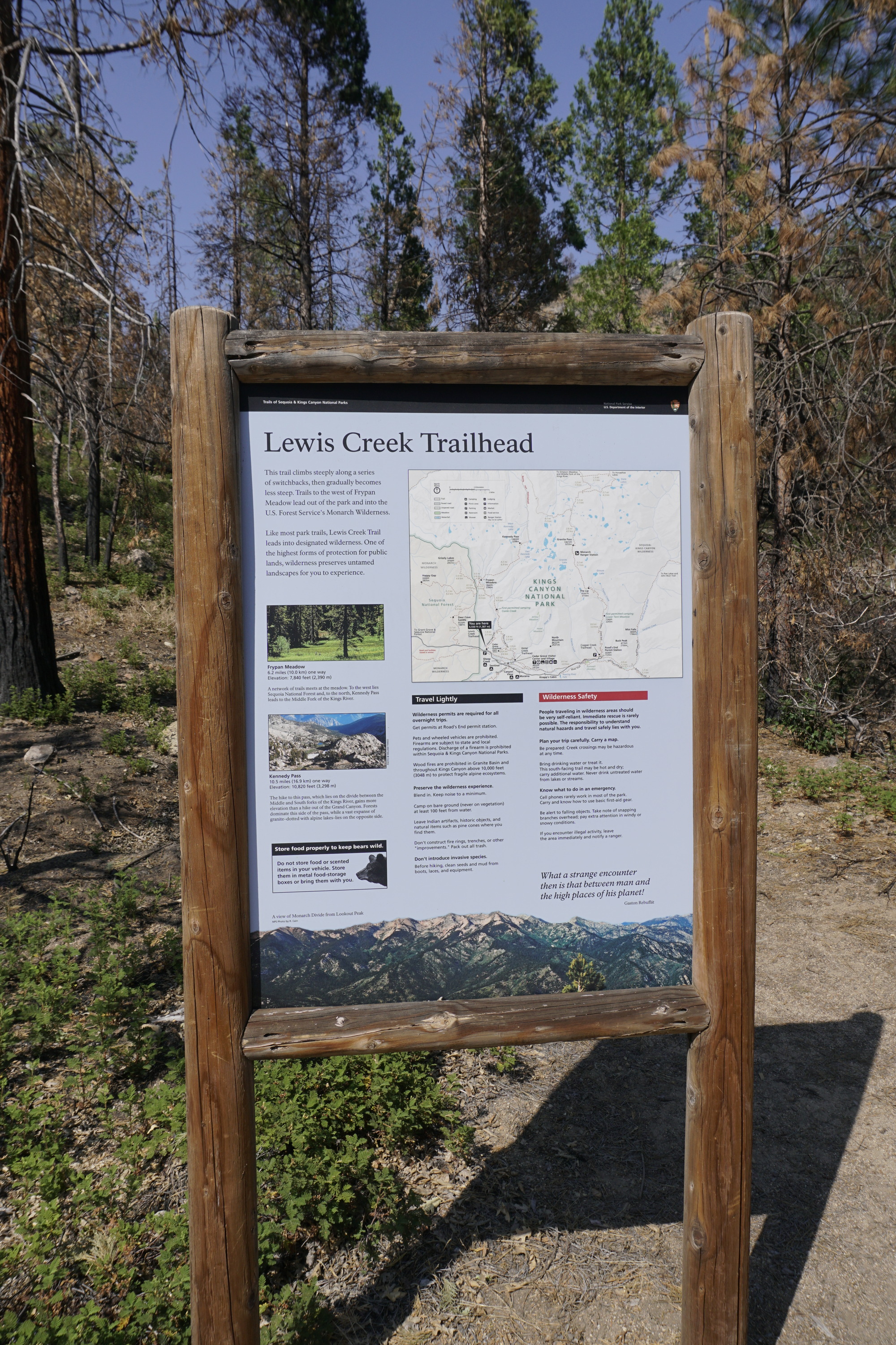 A metal panel with maps on it sits in a wooden frame. Behind the frame are multiple trees. 