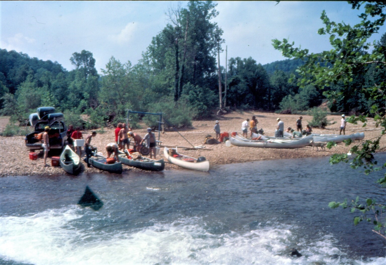 color photo with river in foreground and boats, people and gravel river bank in background.