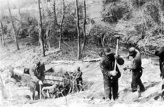A group of men in work clothes hammer and shovel stone from a hillside into a truckbed.