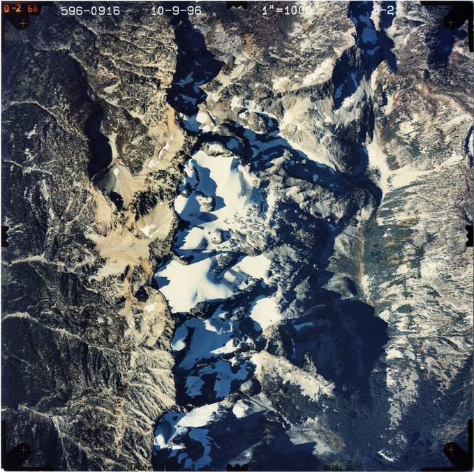 Color aerial photograph of glacier and snowfields on mountain peak with forested valleys below.
