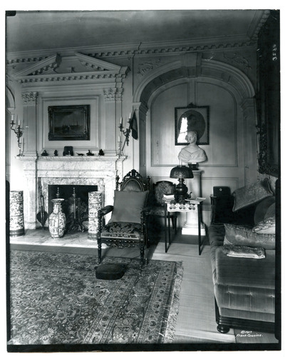 Black and white photograph of 19th century parlor featuring chairs, couches, a bust, and an elaborate fire place.