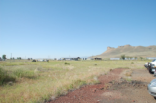 A grass field with distant cars and buildings. A rocky mountain formation is in the background.