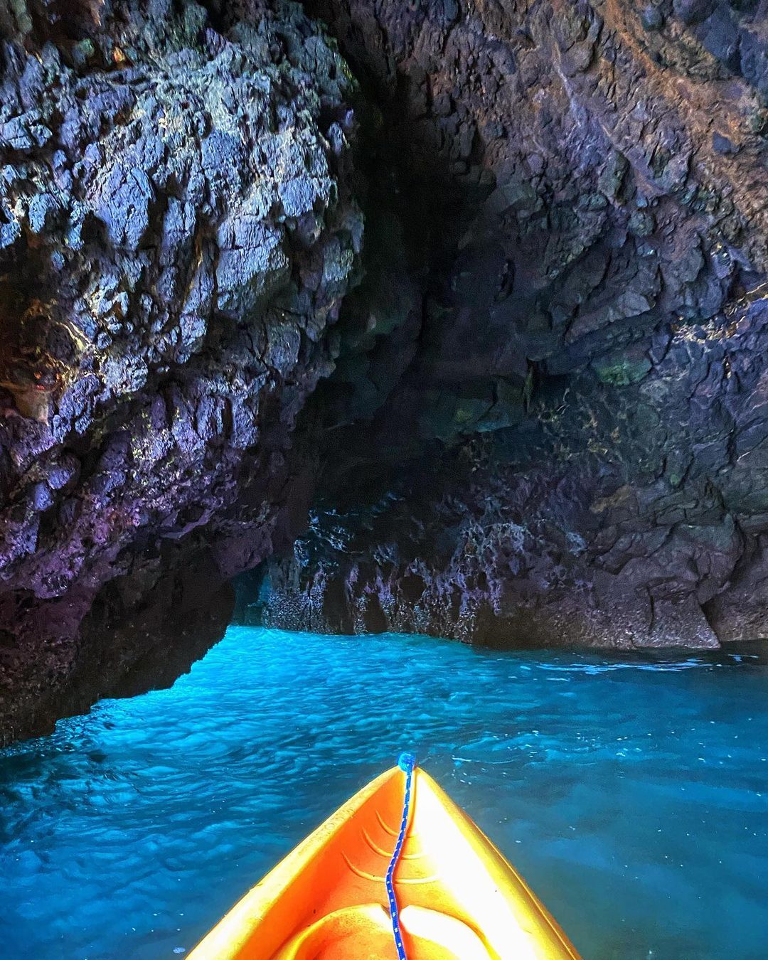 yellow tip of kayak in foreground with purple and green sea cave in background on a emerald green water