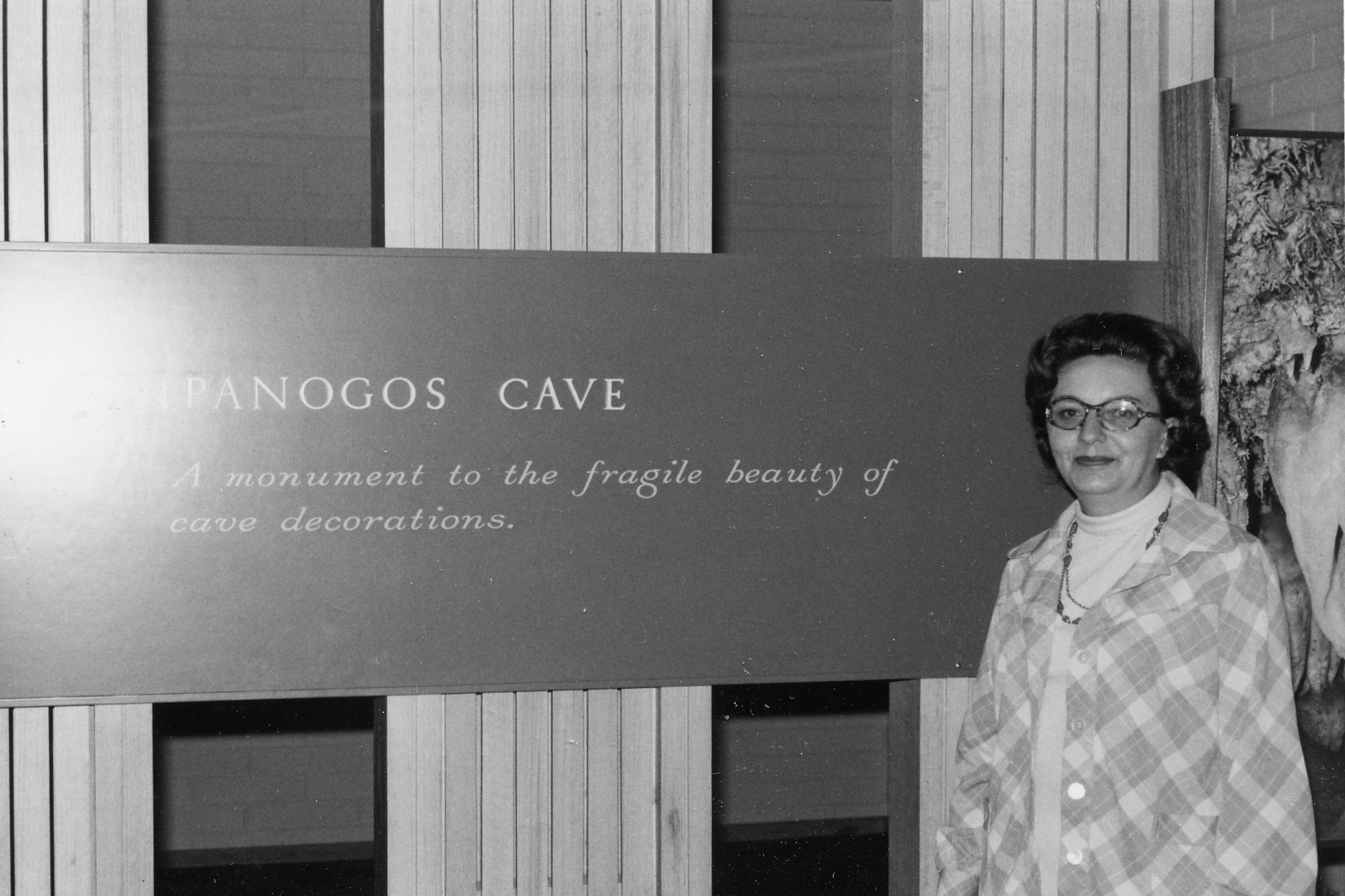 A black and white photo of a woman with short hair and glasses wearing a plaid coat standing in front of a sign that reads: Timpanogos Cave A monument to the fragile beauty of cave decorations.