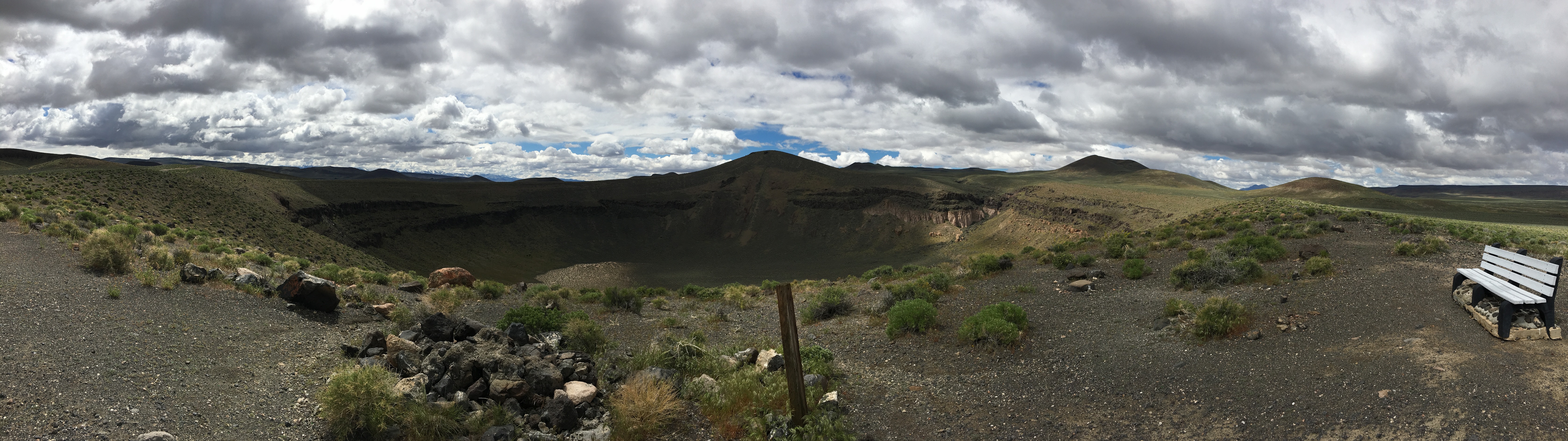 A panorama of Lunar Crater National Natural Landmark. The crater sinks into the earth and there are volcanic cones surrounding the crater. On the rim of the crater, there is a bench for visitors to sit and enjoy the view.